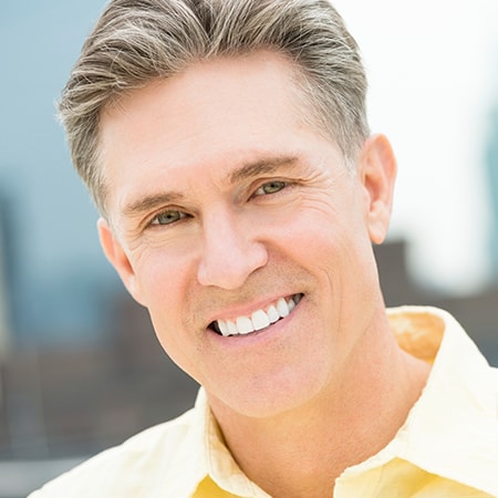 Experience the joy of a renewed smile A mature man smiling while wearing a yellow shirt and one of our dental bridges